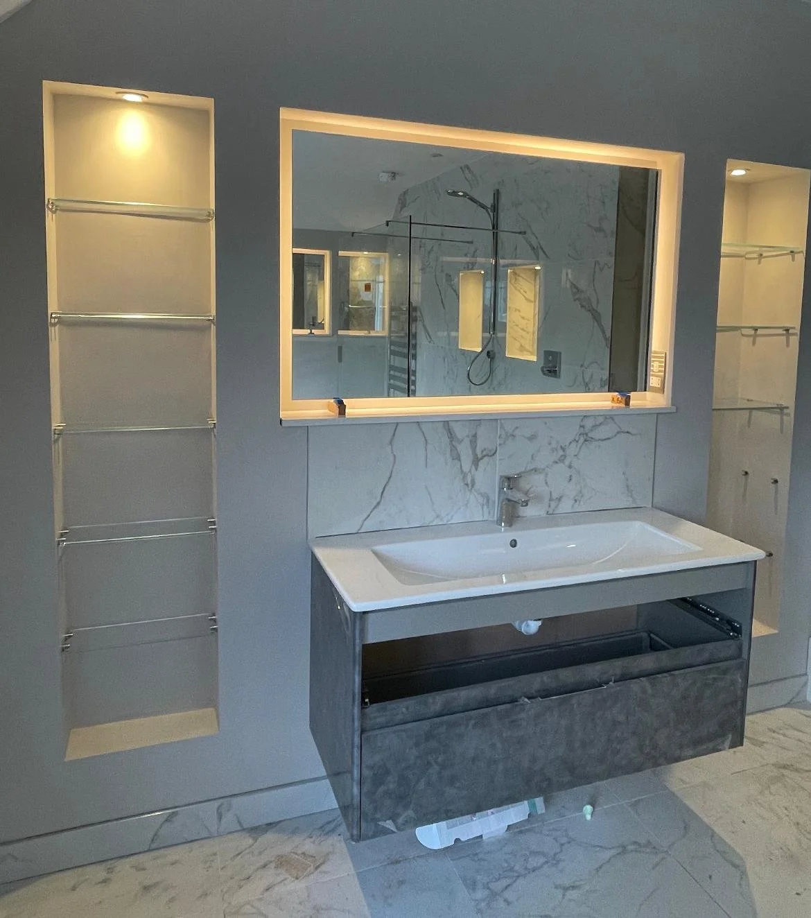 Large bespoke bathroom wall mirror installed above marble vanity with illuminated recessed shelving and glass display shelves in a luxury Surrey bathroom renovation.