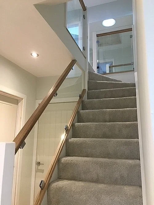 Indoor staircase with gray carpet, wooden handrail, glass panels, leading to a second-floor hallway with a glass railing and ceiling lights.
