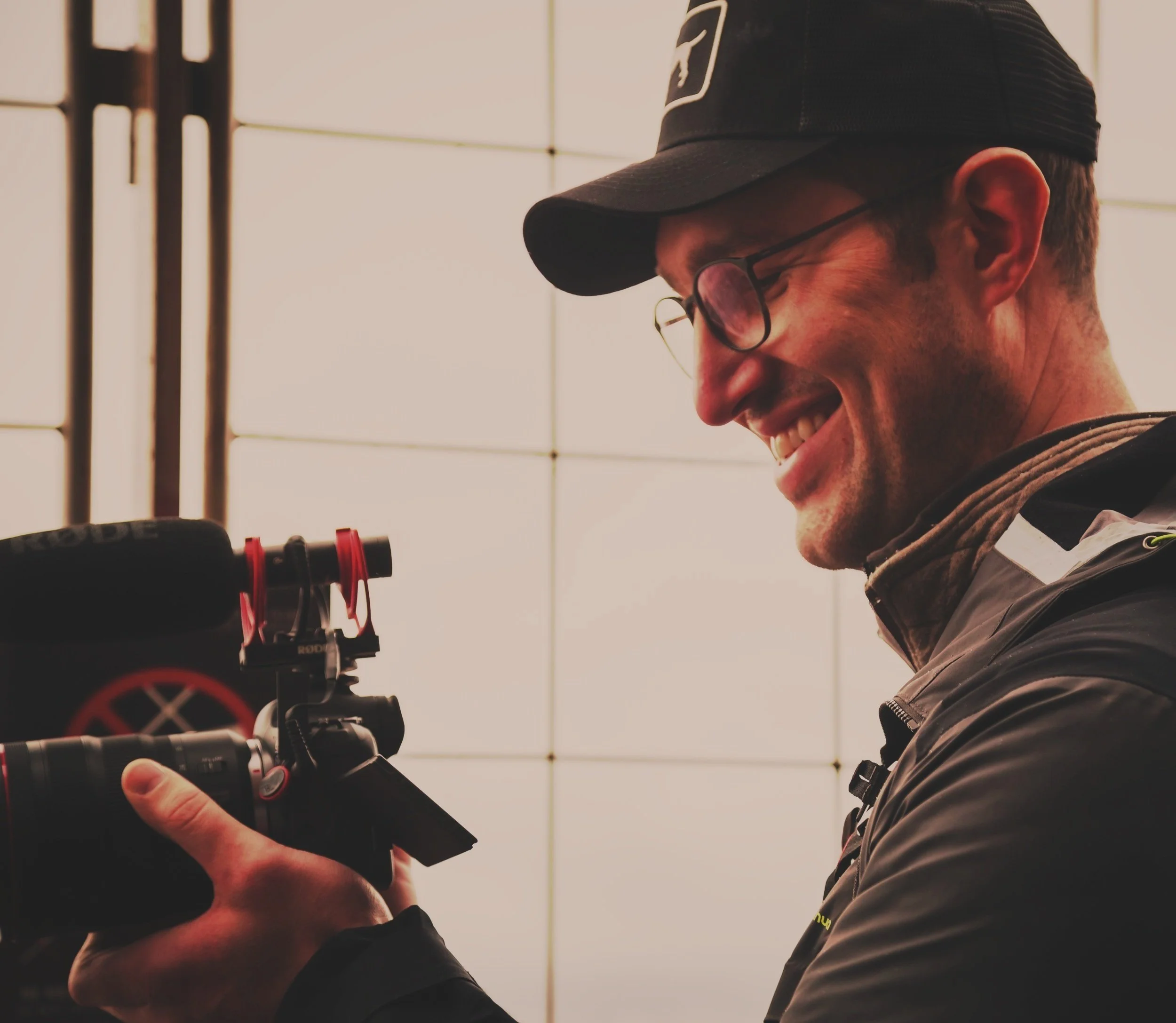Smiling person in glasses and cap holding camera with mounted microphone in indoor setting.