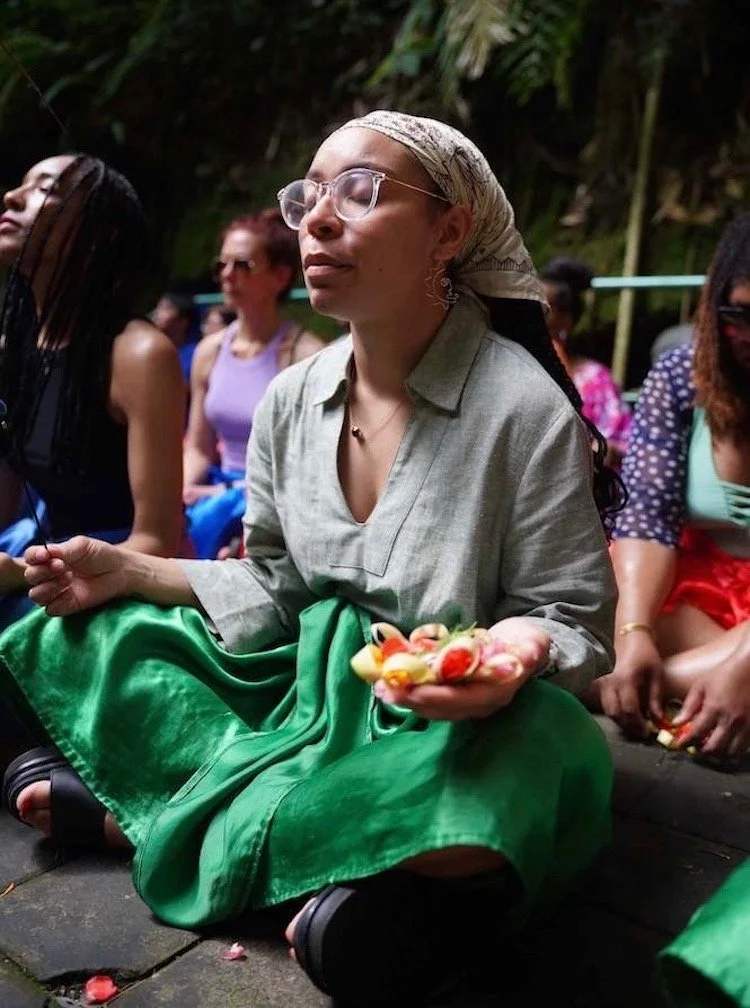 Woman meditating in Bali with eyes closed, gently exhaling. Holding an incense stick and a traditional Balinese offering, wearing a green dress and silk covering copy