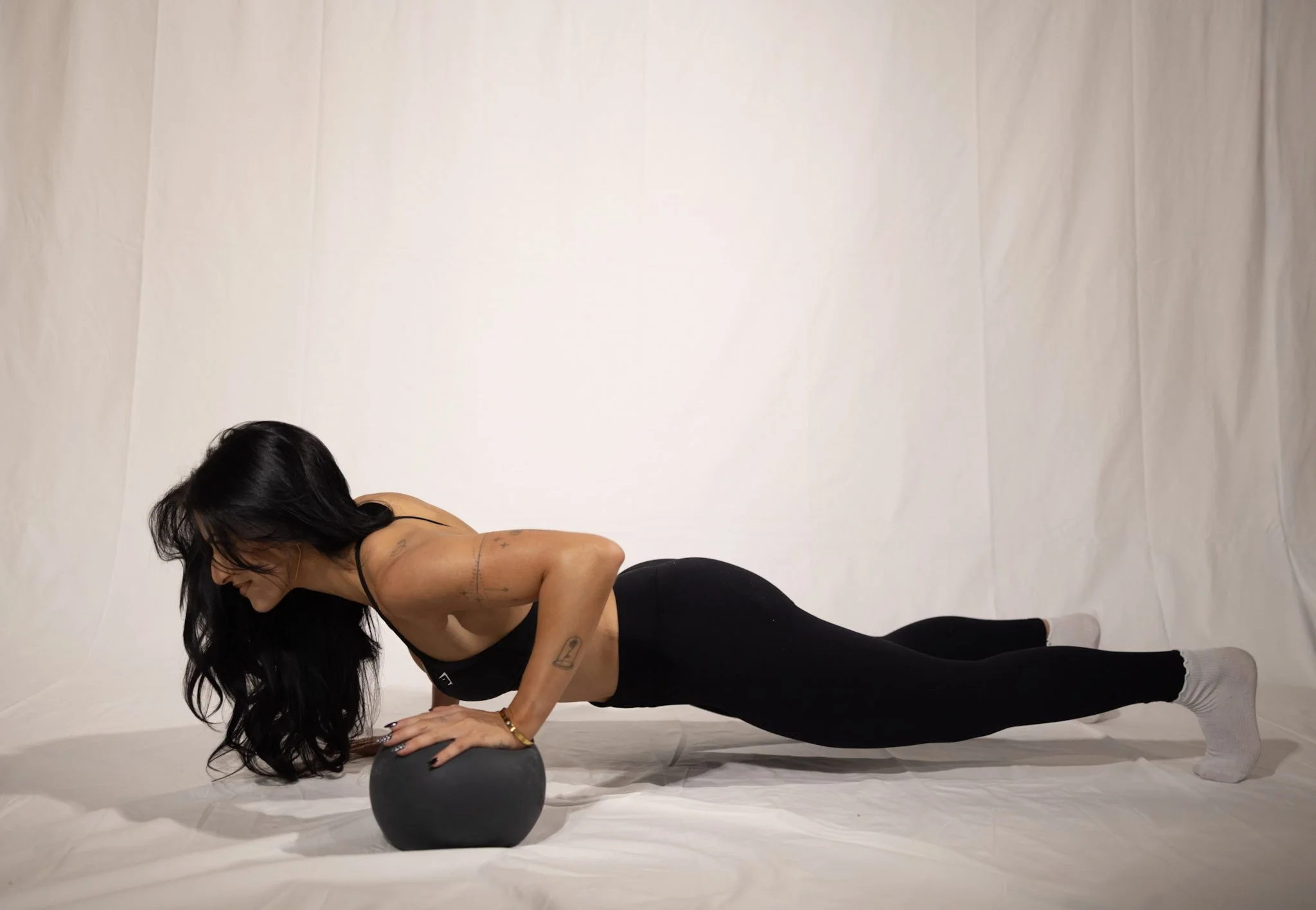 Woman in black sports bra and leggings doing a push-up with hands on a black exercise ball, on a white background.