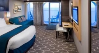 A hotel room with a large bed, blue and white bedding, a TV on the wall, a small desk, and a balcony with a view of the ocean.