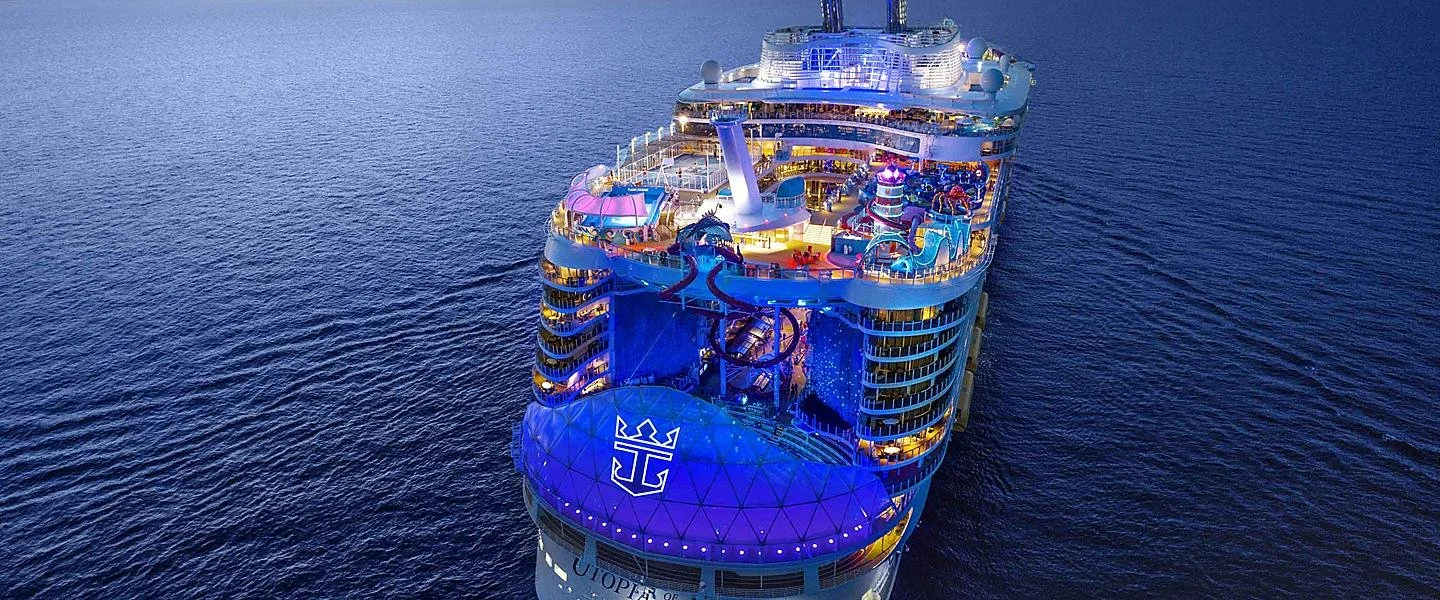 An aerial view of a large cruise ship sailing at sea, illuminated with colorful lights around the decks.