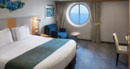 Hotel room with a large bed, blue armchair, round window showing ocean view, and a small table.