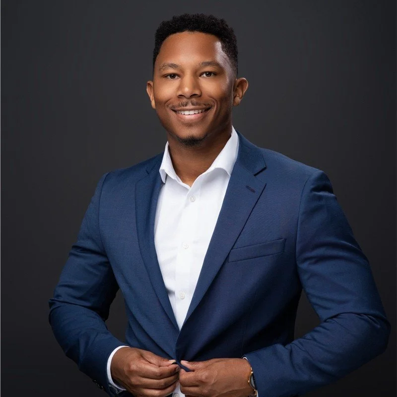 A man in a blue suit and white shirt smiling, standing against a dark gray background.