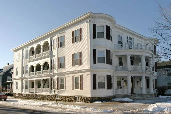 A large white multi-story residential building with rounded balconies and arched openings on the upper floors, situated in a snowy environment.