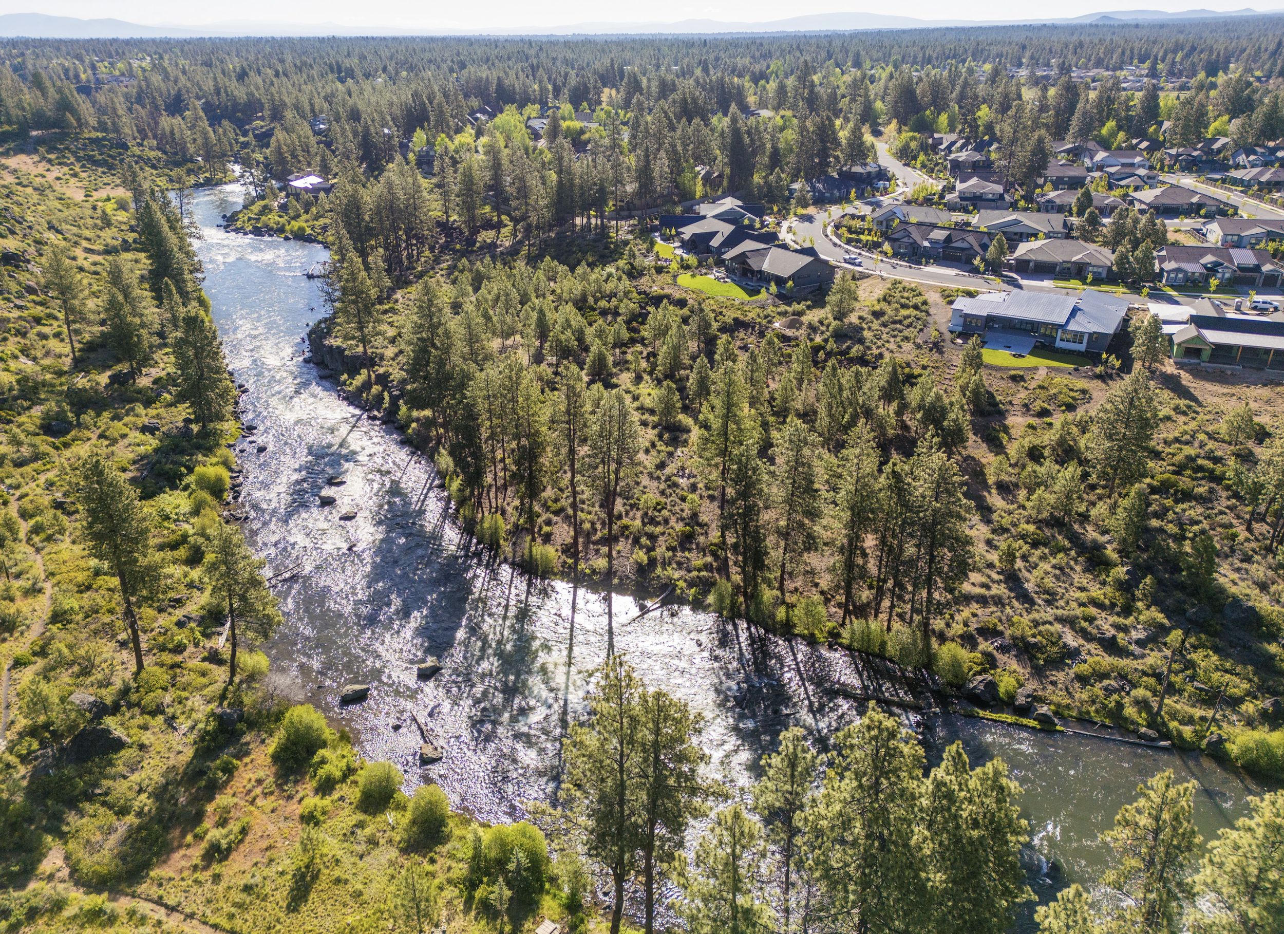 Aerial view of a river flowing through a forested area with residential homes in the background