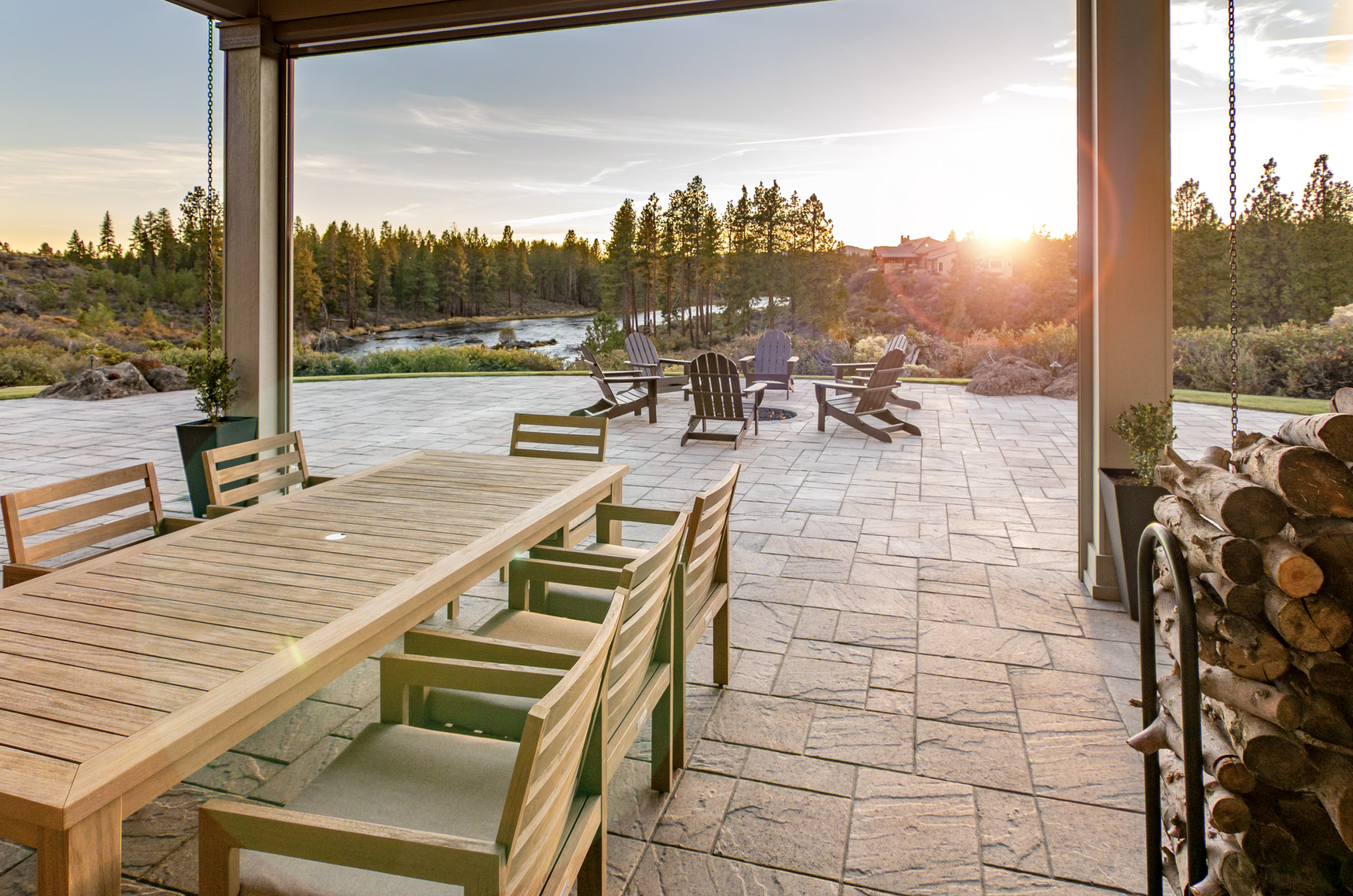 Outdoor patio with wooden dining table, chairs, fire pit, and Adirondack chairs. Surrounded by trees and a river, with sunset in the background.