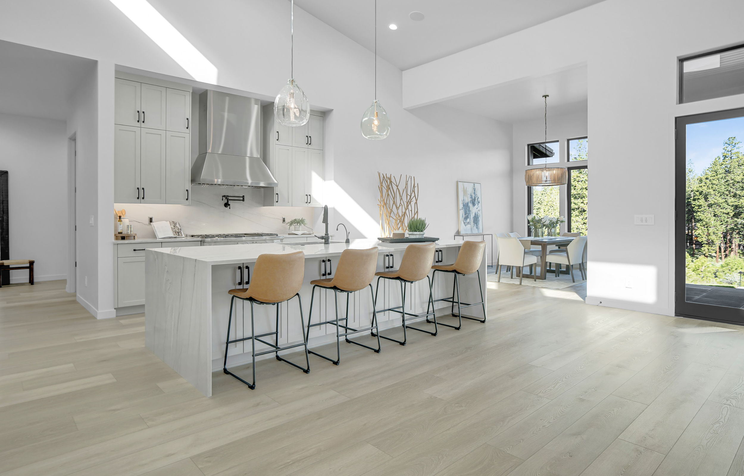 Modern kitchen with white cabinets, center island, brown bar stools, and pendant lights. Dining area with table and chairs, large windows overlooking trees.