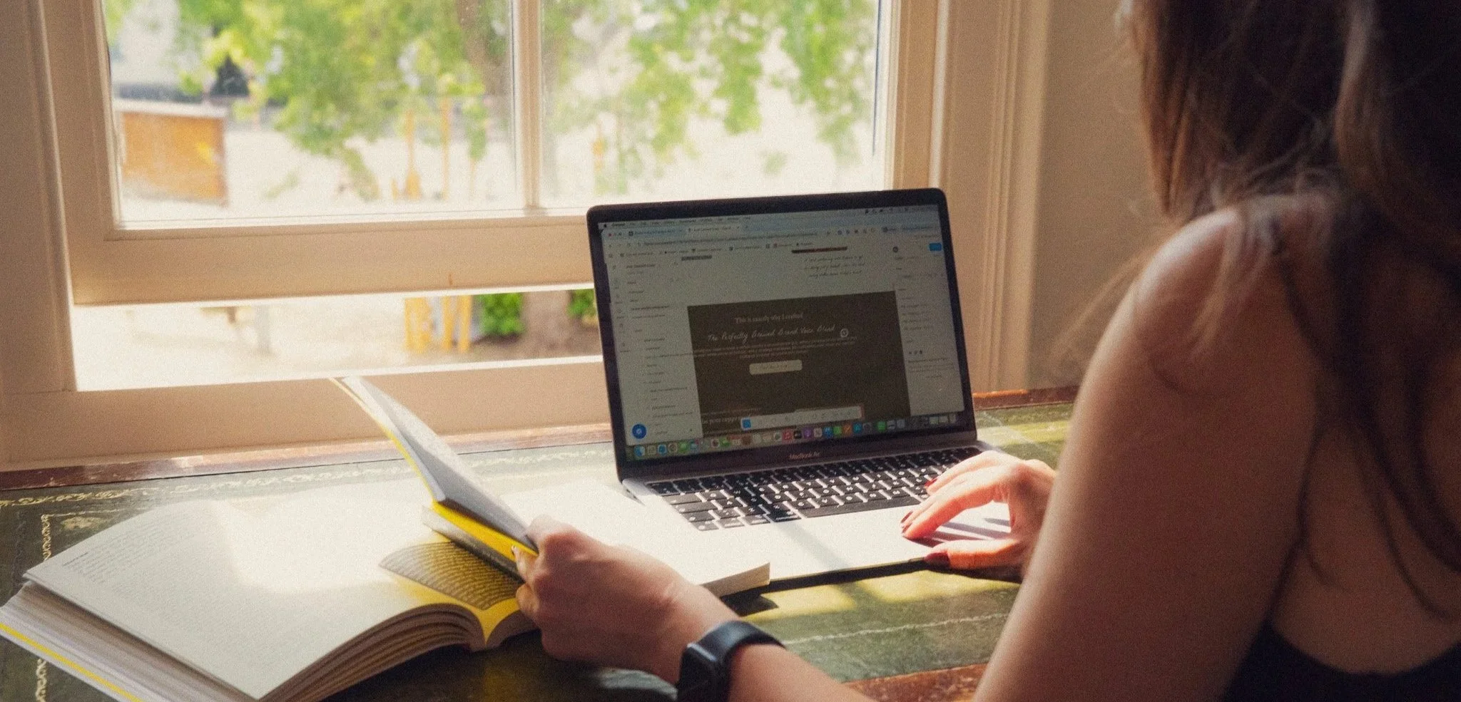 Hols, brand writer sat at a desk typing on a laptop with a hand on a book in front of a window