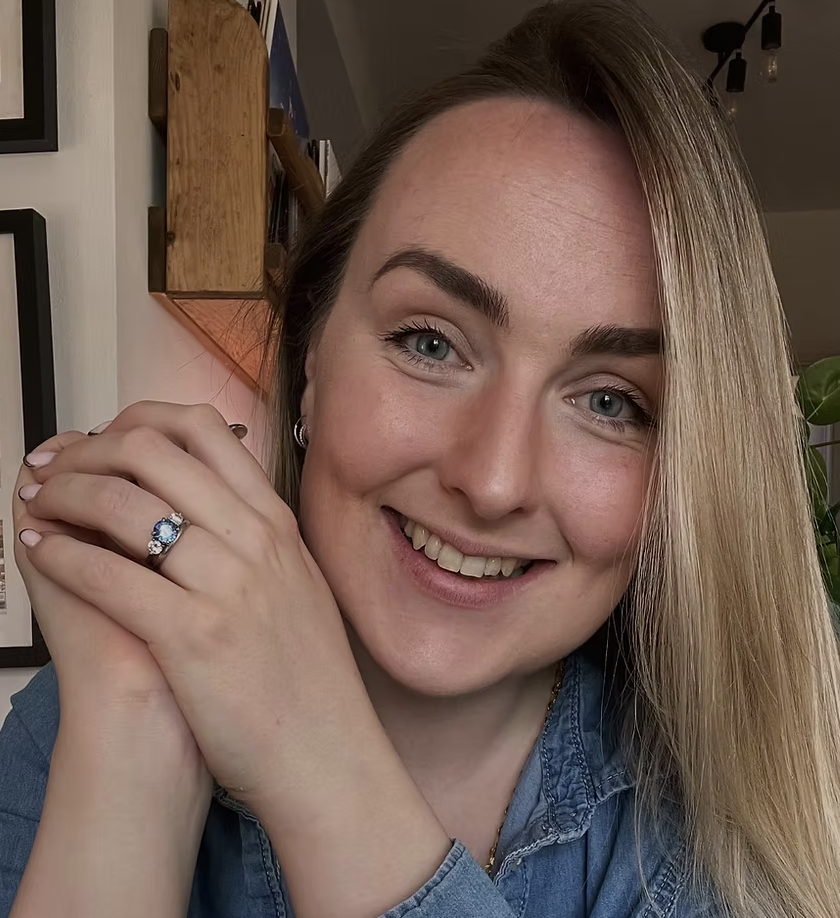 Close-up of a smiling woman with blonde hair, wearing a blue shirt, with her hands near her face, showing an engagement ring with a blue gemstone.