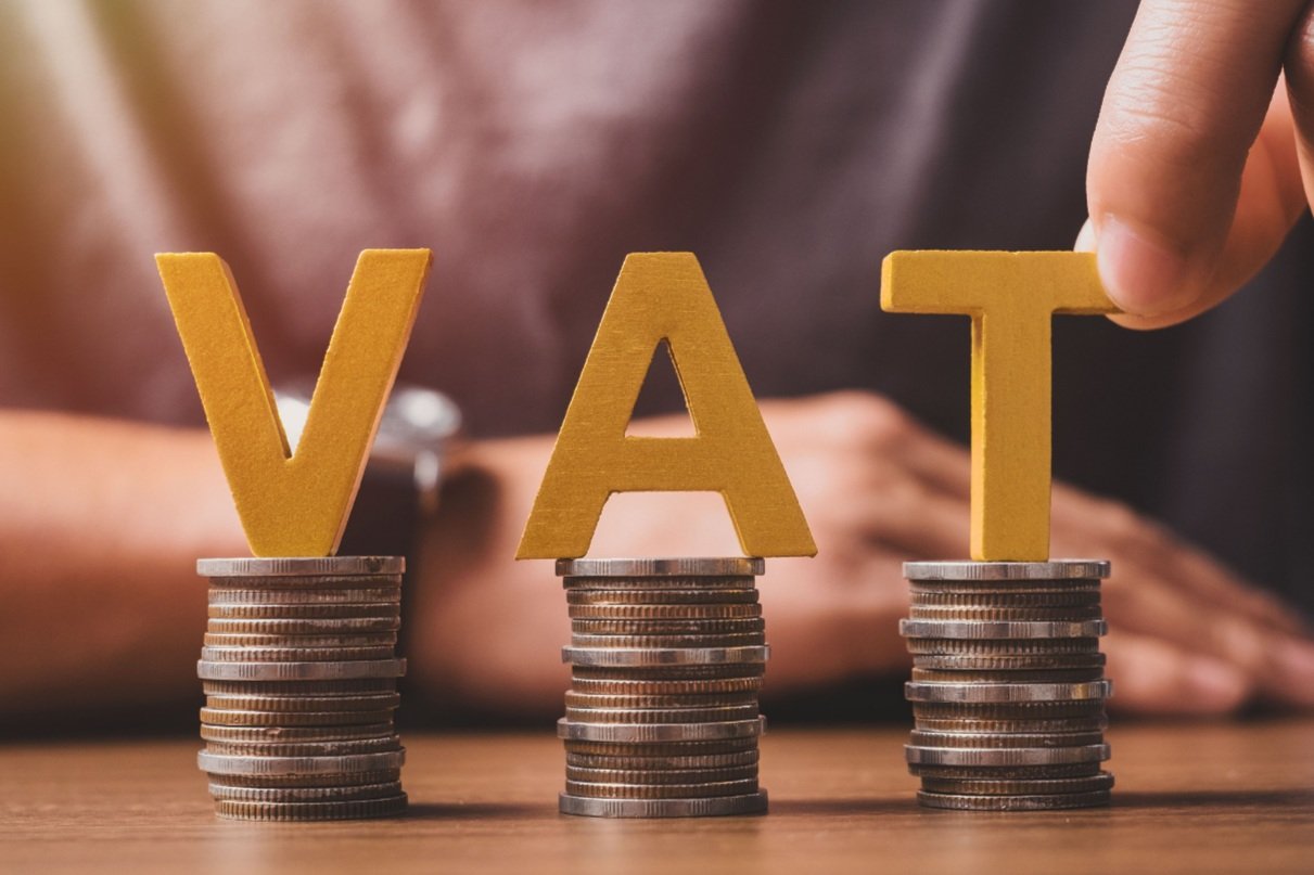 Stacked coins with large yellow letters "V, A, T" on each stack, representing "Value Added Tax".