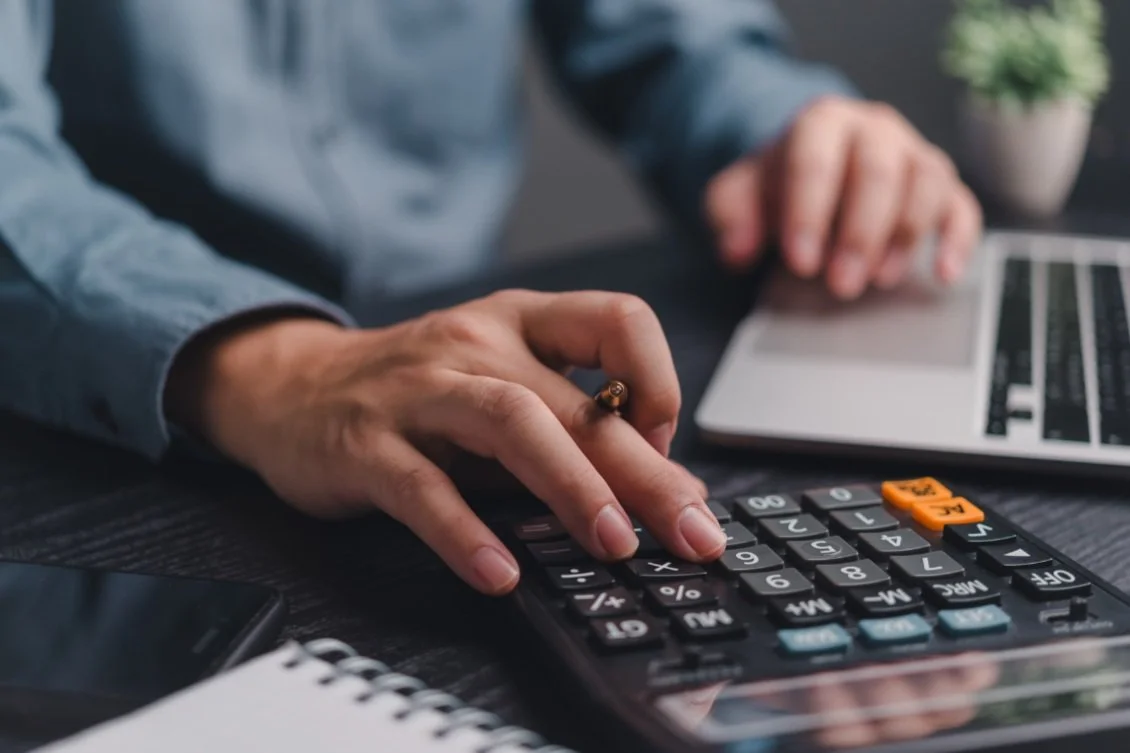 Person using a calculator and laptop at a desk with a notebook and smartphone visible.