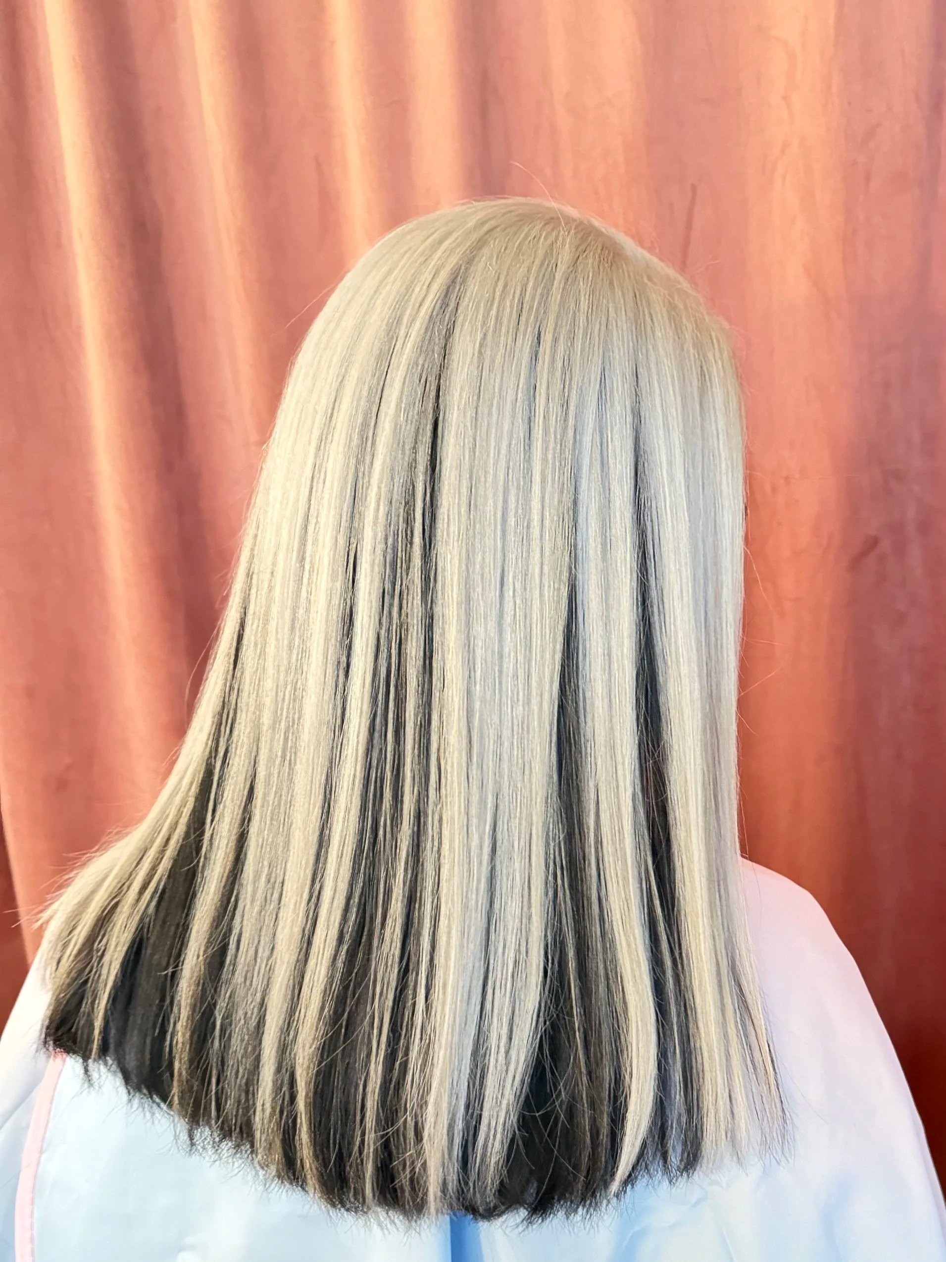 Woman with long, wavy, blonde hair facing away from the camera, standing in front of pink curtains.