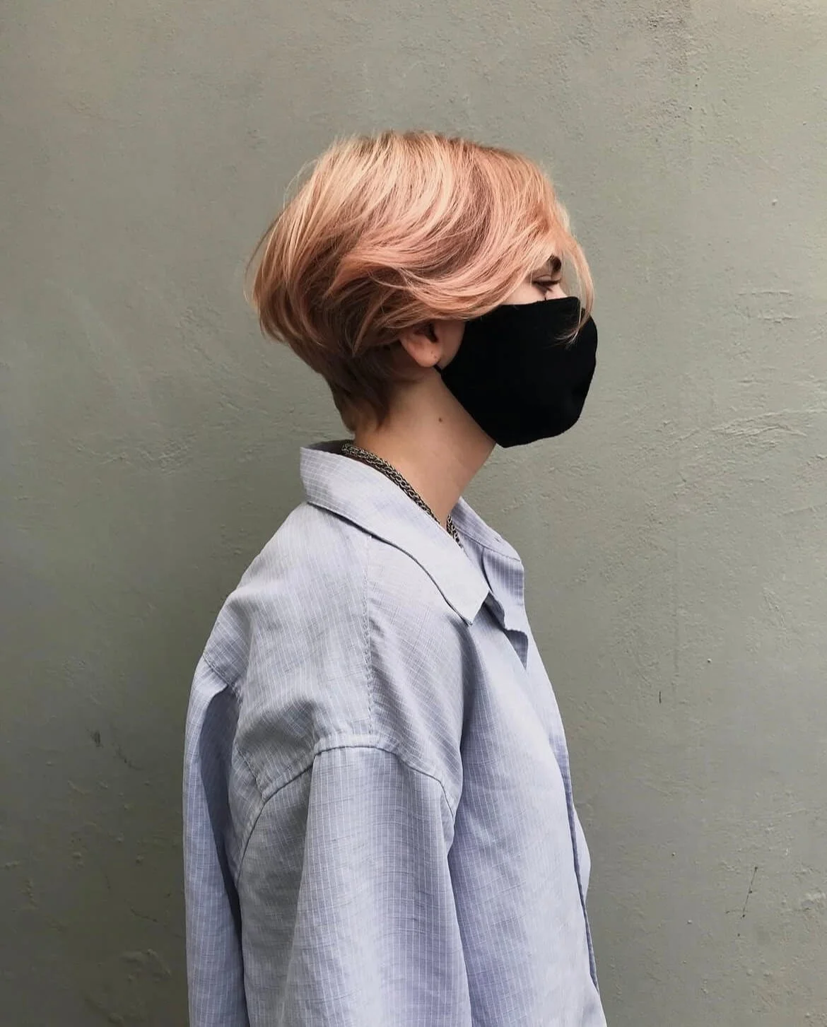 Side profile of a person with short, layered, light pink hair wearing a black face mask and a light blue button-up shirt.