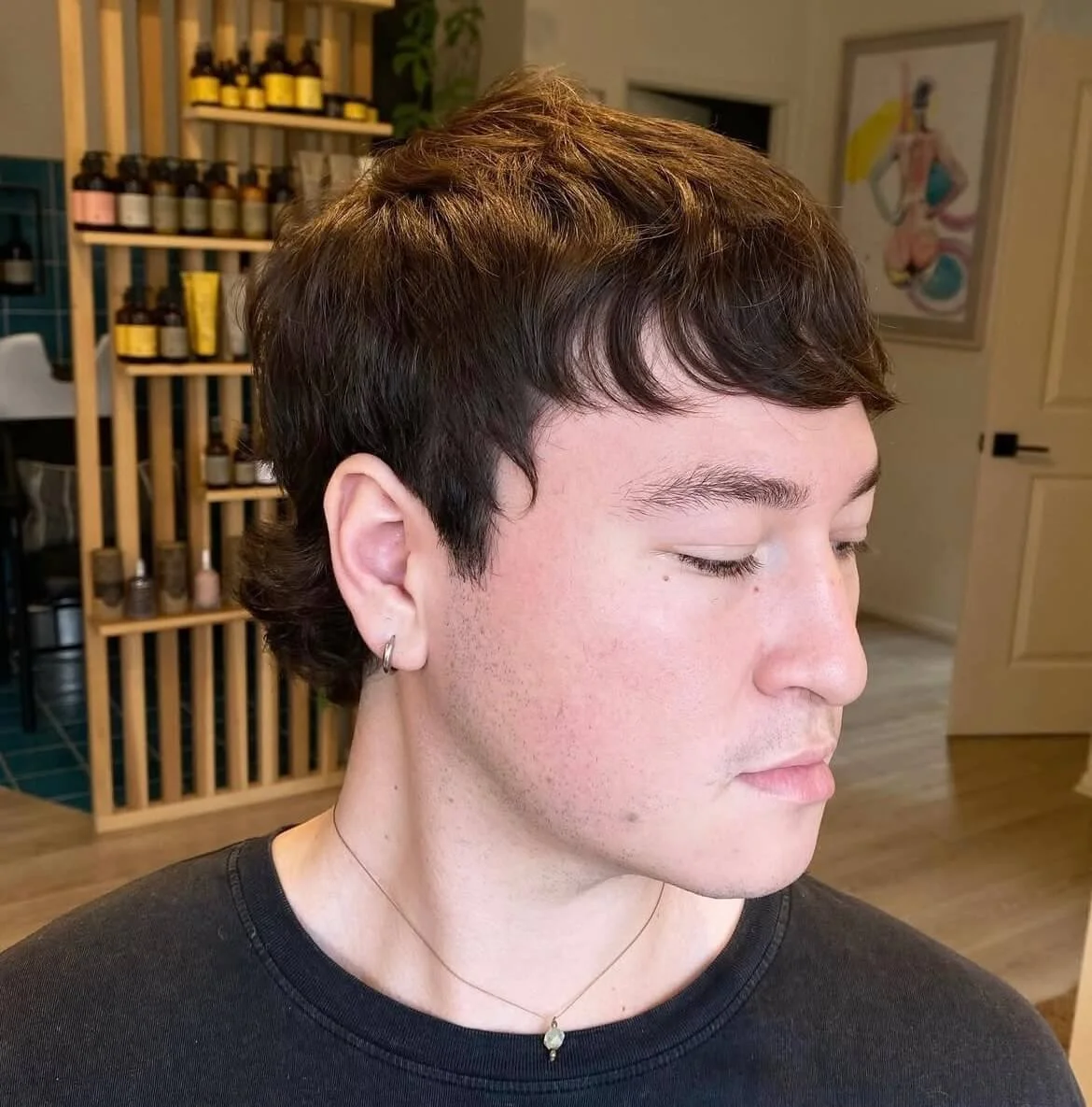 Person with short dark hair, wearing an earring and necklace, looking down, in a modern room with shelves and a painting.