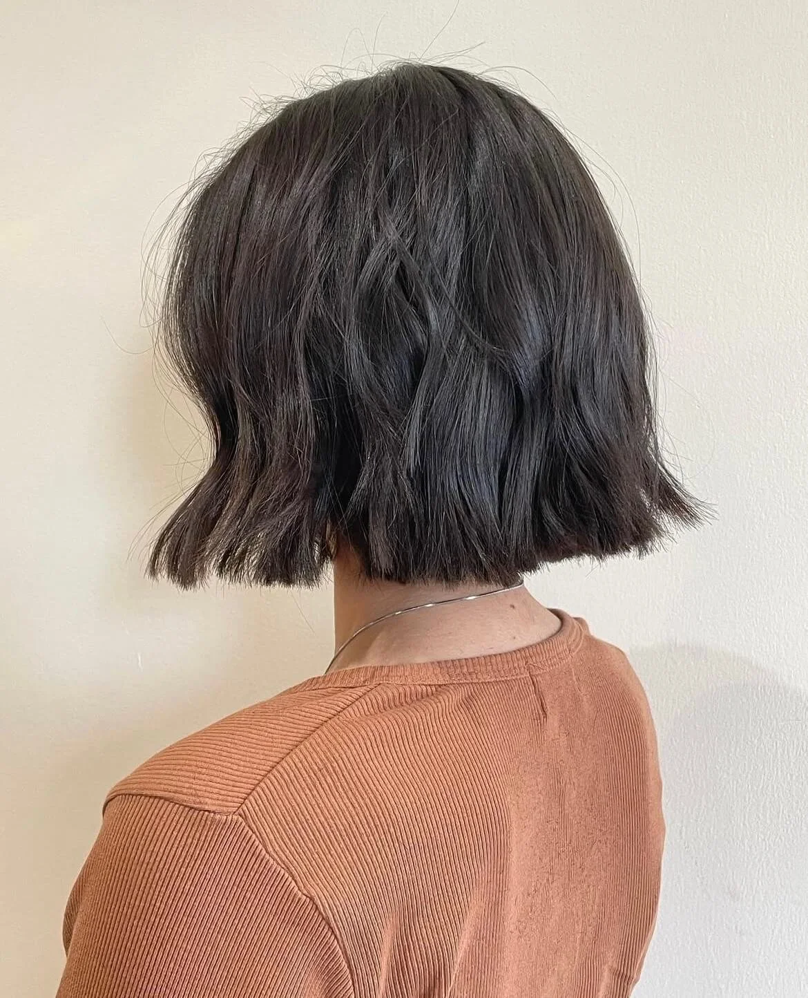 Back view of a person with a short, wavy brown bob haircut, wearing a brown ribbed shirt.