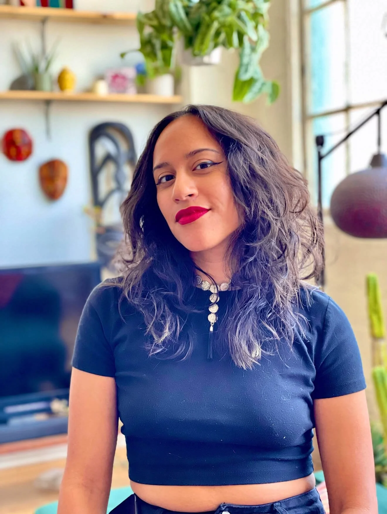 Woman with curly hair wearing a black crop top, standing indoors with a plant and decorative elements in the background.