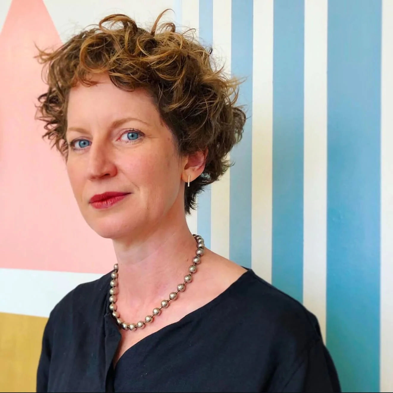 Person with short curly hair wearing a pearl necklace and dark top, standing in front of a colorful striped wall.