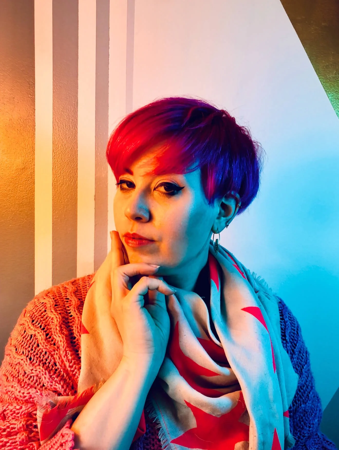 Person with bright pink hair and a contemplative expression, wearing a knitted sweater and patterned scarf, with colorful striped wall background.