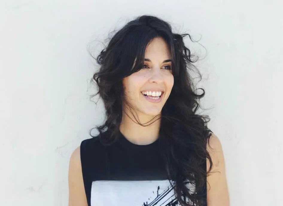 Smiling person with long, curly hair standing against a light-colored background.