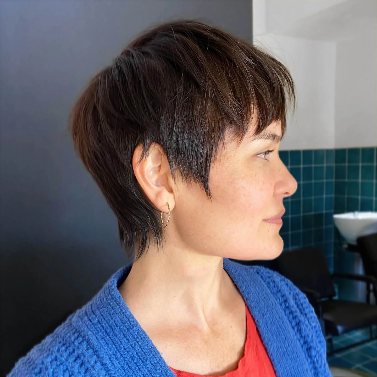 A person with a short, textured pixie cut in warm brown tones, wearing a blue cardigan over an orange top, shown in profile in a salon setting.