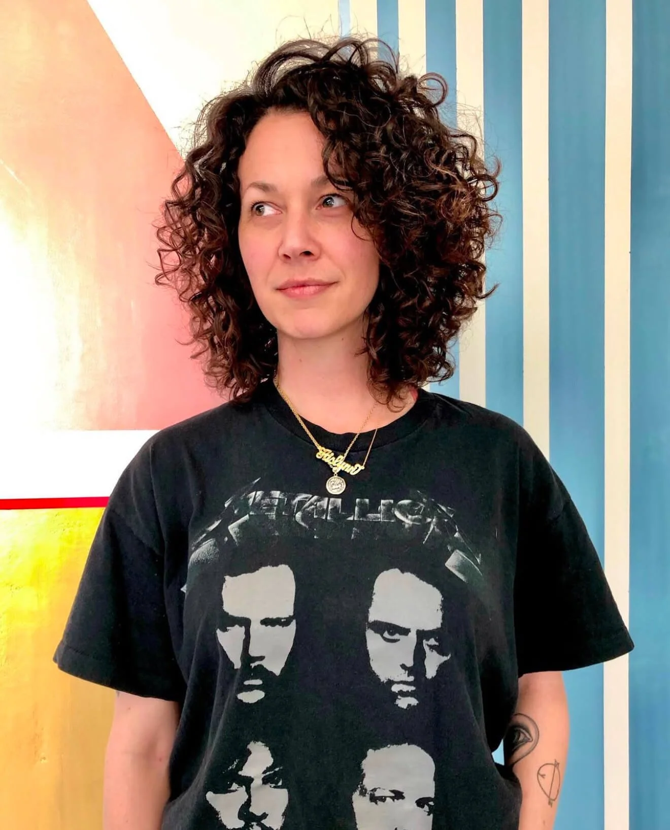 Person with curly hair wearing a Metallica t-shirt, standing in front of a colorful, geometric background.