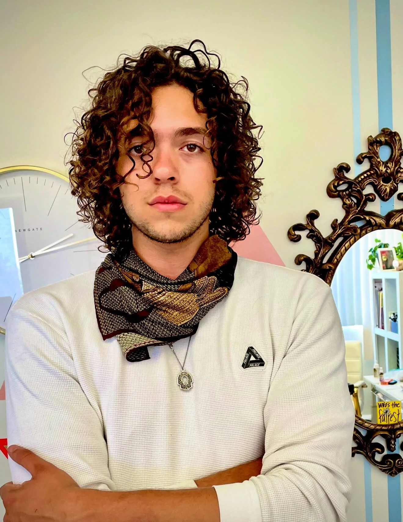 Person with curly hair wearing a patterned scarf and white long-sleeve shirt, standing in front of a decorative wall clock and ornate mirror, with crossed arms and a pendant necklace.