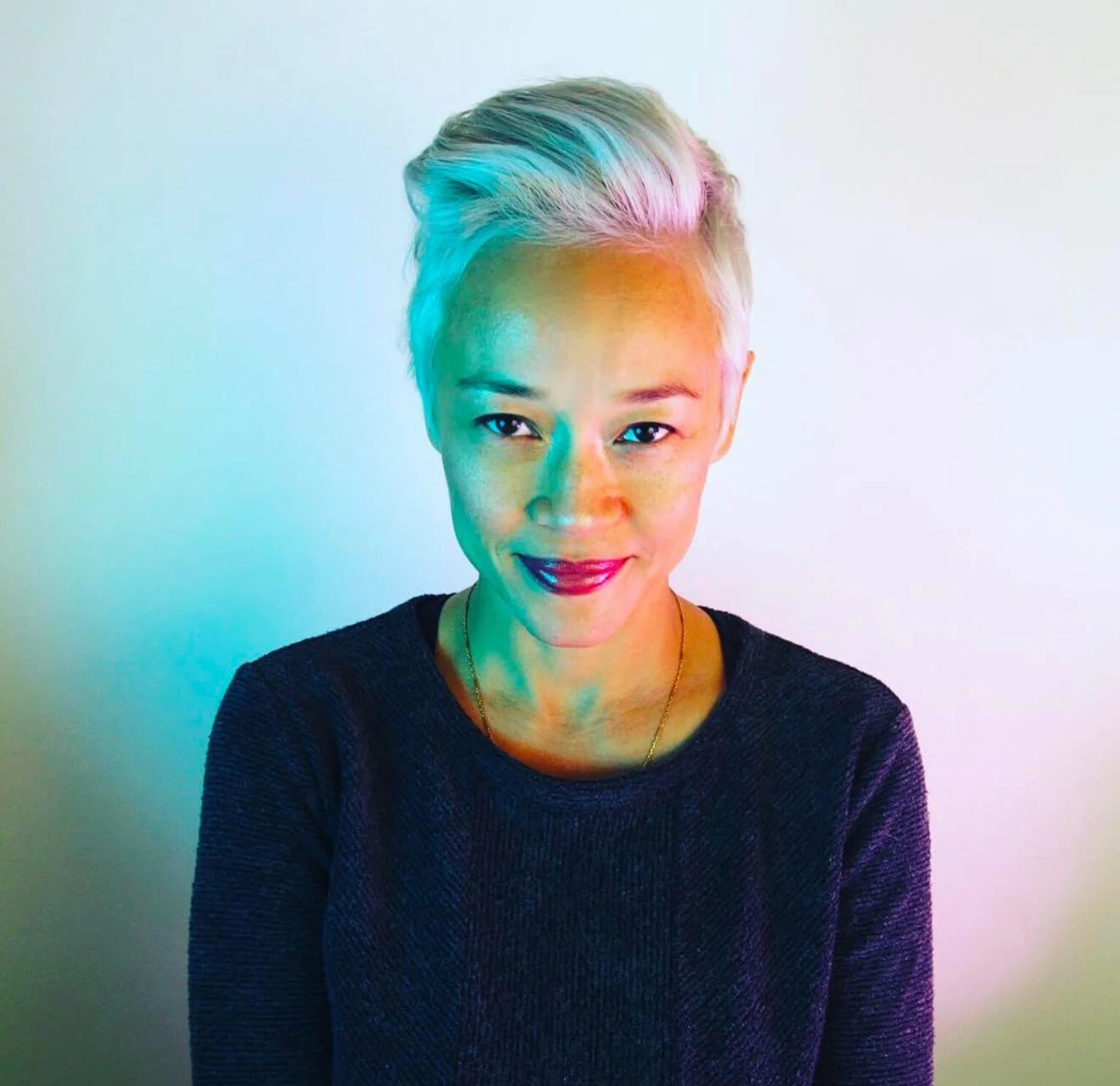 Person with short white hair and a blue sweater, smiling with colorful lighting on face.