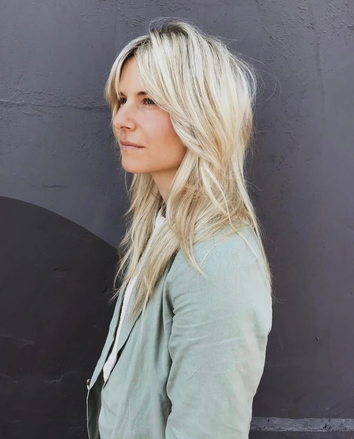 Person with long blonde hair wearing a light-colored jacket, standing against a dark gray wall.