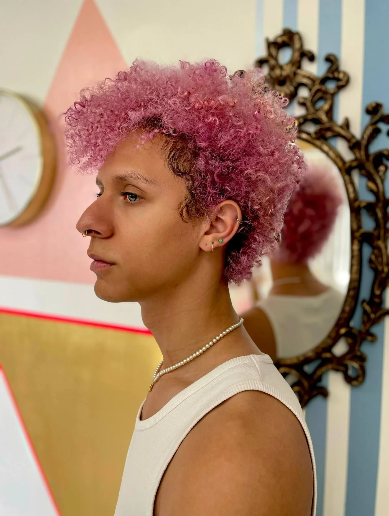 Person with pink curly hair in profile, wearing a white tank top and a pearl necklace, standing in front of a mirror with an ornate frame. Background includes a clock and colorful wall design.