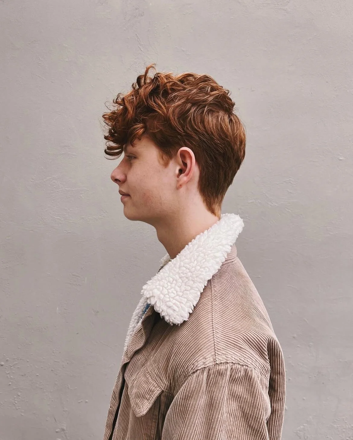 Profile view of a person with curly red hair wearing a brown corduroy jacket with a fleece collar against a neutral background.