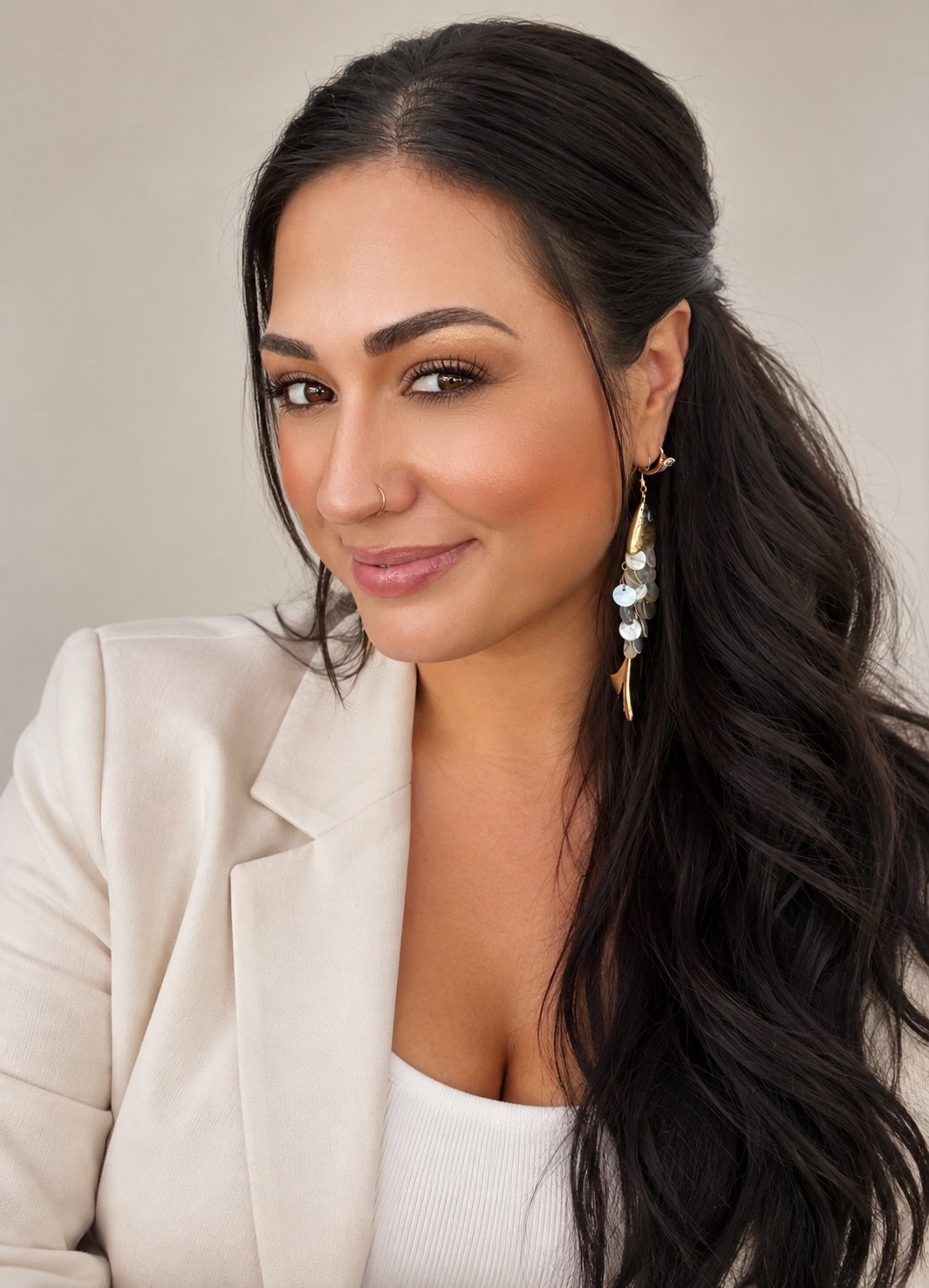 Close-up of a smiling woman with long dark wavy hair, wearing a white blazer and large earrings.