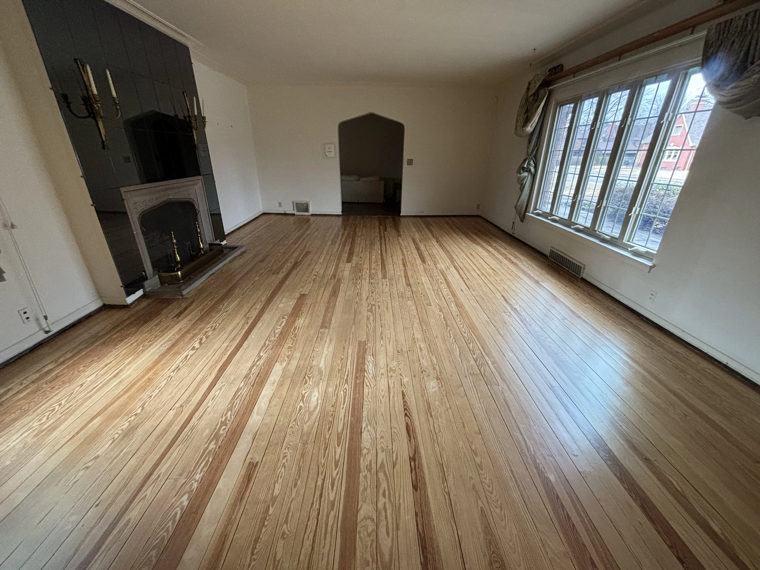 Empty living room with hardwood floors, a fireplace with a black mantel, large window with drapes, and an archway leading to another room.