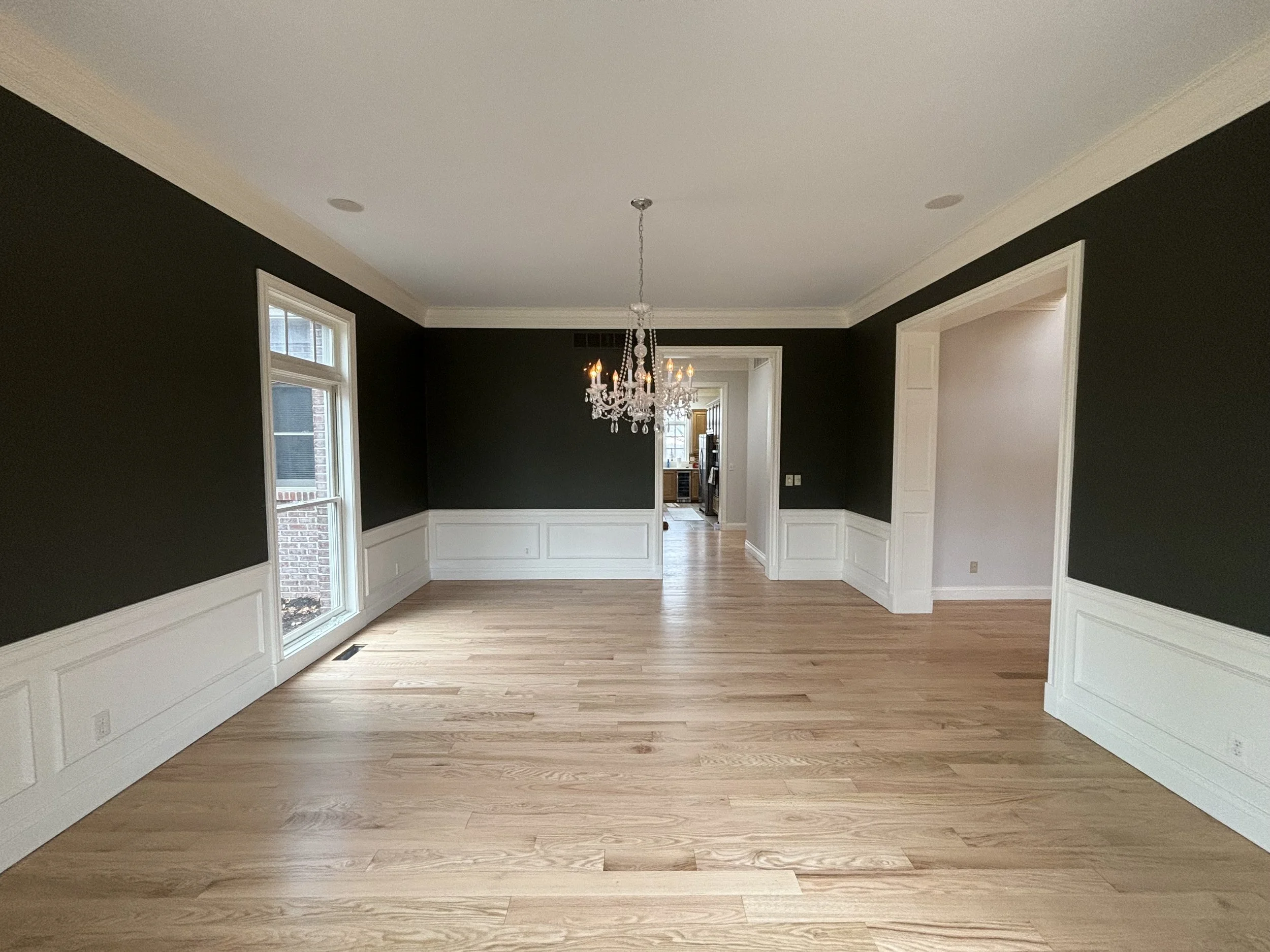 Empty living room with dark green walls, white trim, large windows, hardwood floors, and a chandelier.