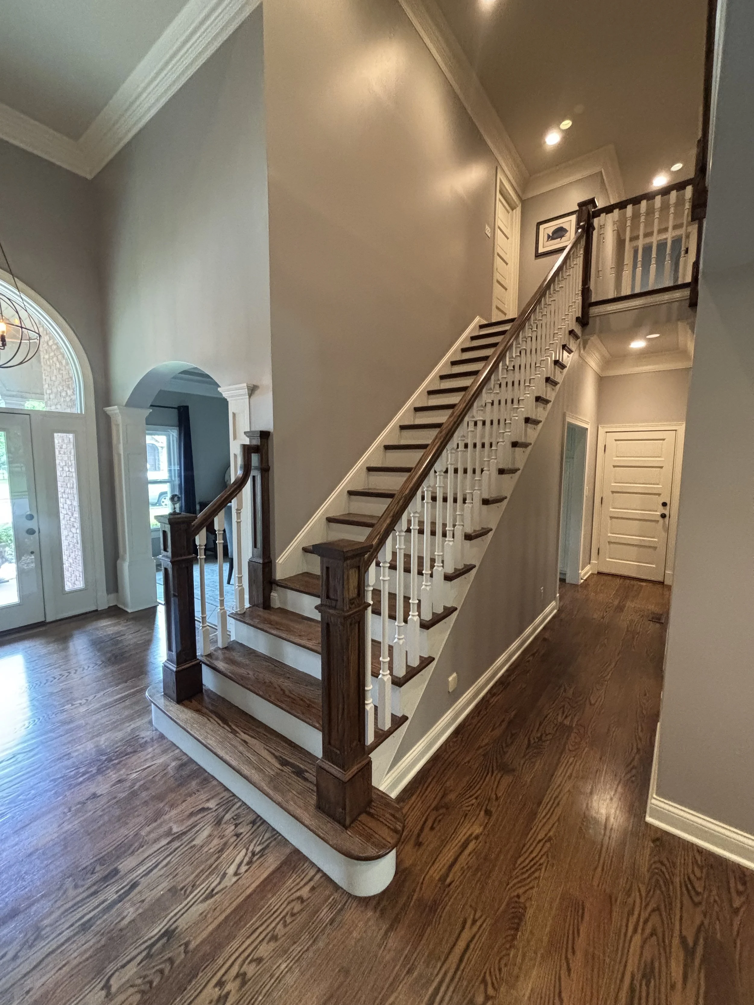 Interior view of a house entryway with a staircase featuring dark wood handrails and white spindles, hardwood flooring, and a front door with glass panels. There's an archway and a framed picture on the wall at the top of the stairs.