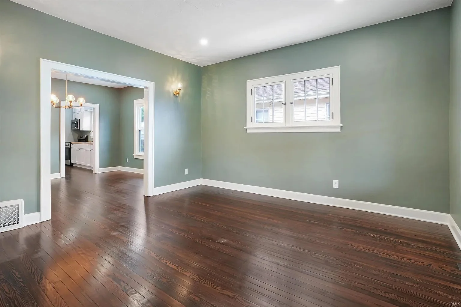 Empty living room with green walls, hardwood floors, a window with blinds, and a doorway leading to a kitchen.