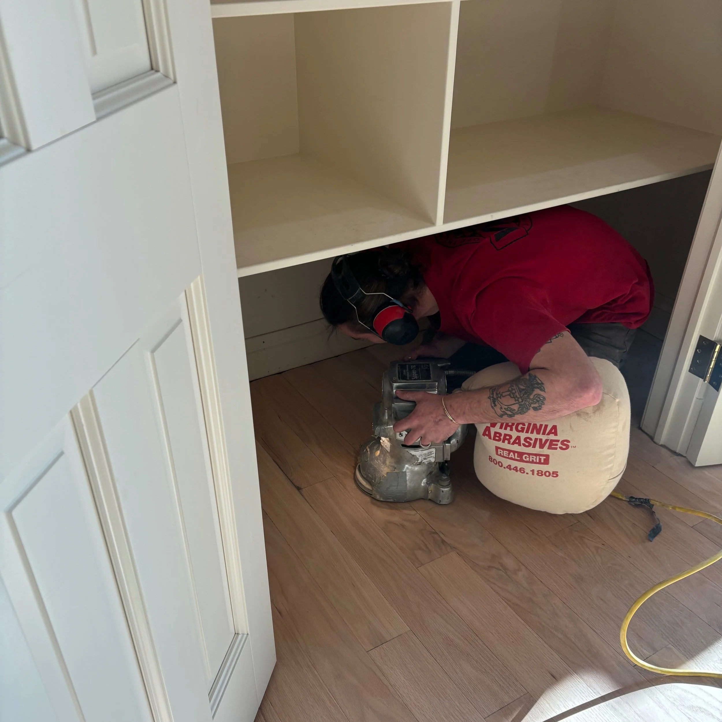 A person working on hardwood flooring under a shelf, using a power tool, wearing ear protection, a red shirt, and sitting on a bag of abrasives.