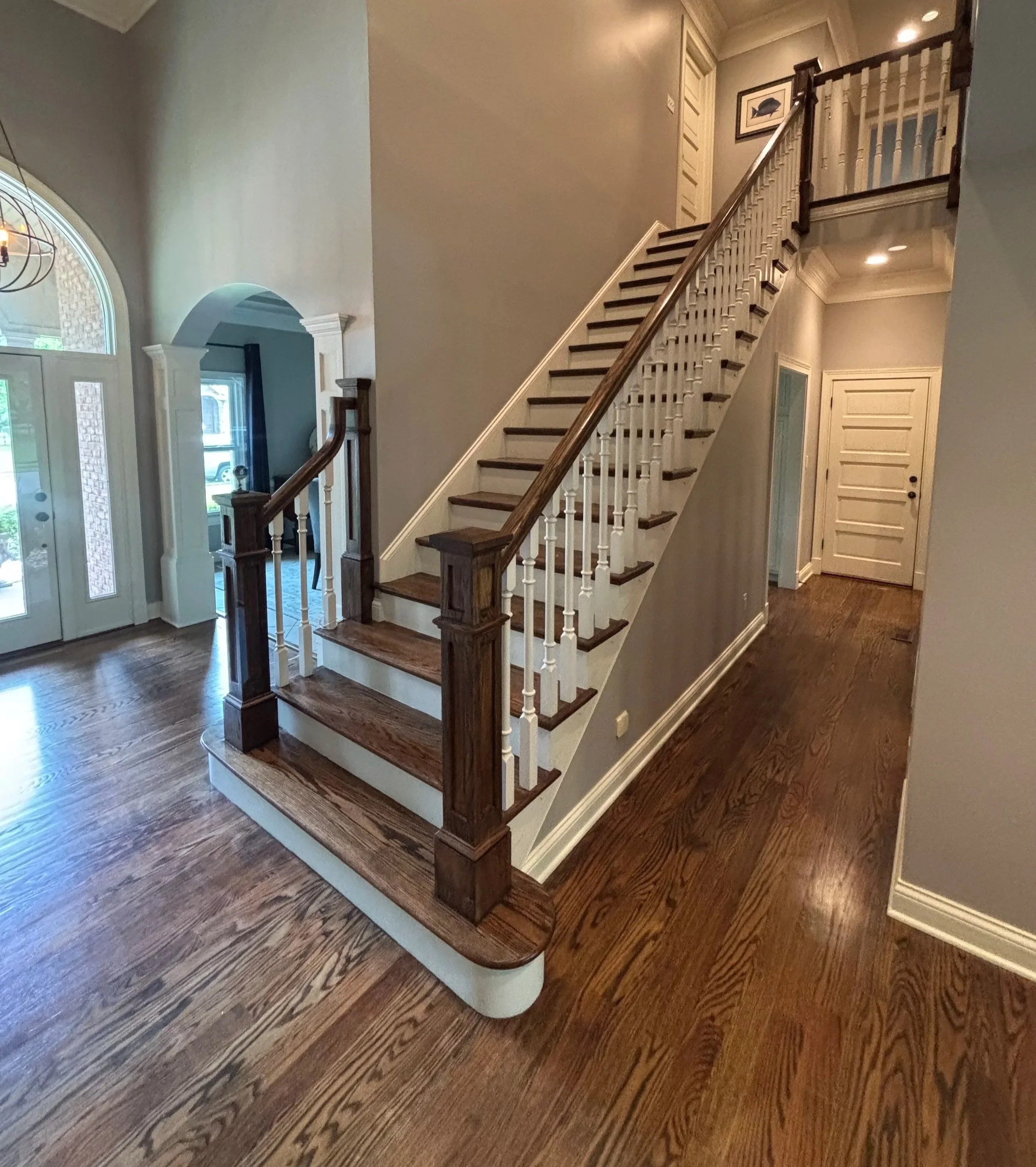 Interior view of a house entryway showing wooden stairs with a dark wood handrail and white spindles, hardwood flooring, and a front door with glass panels.