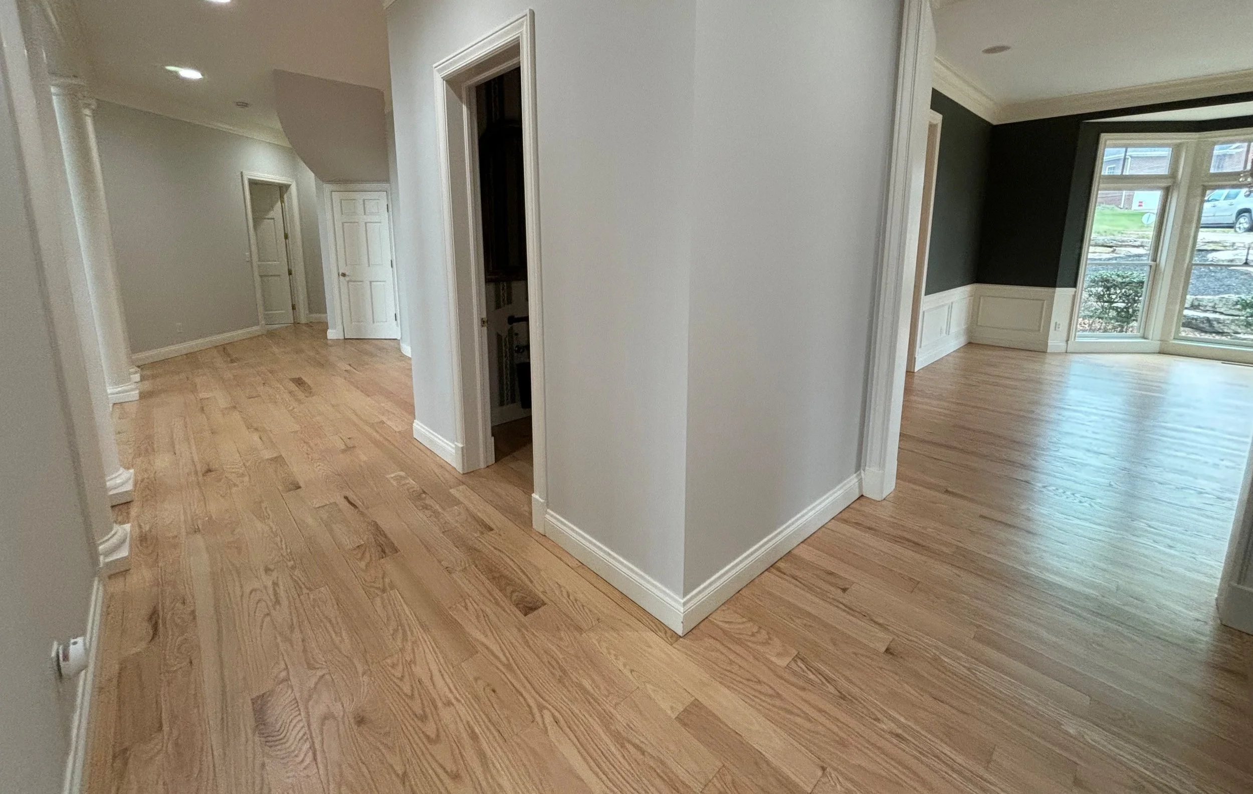 Interior view of a house with light wood flooring, white walls, and multiple rooms visible, including a sunlit room with large windows and a dark accent wall.