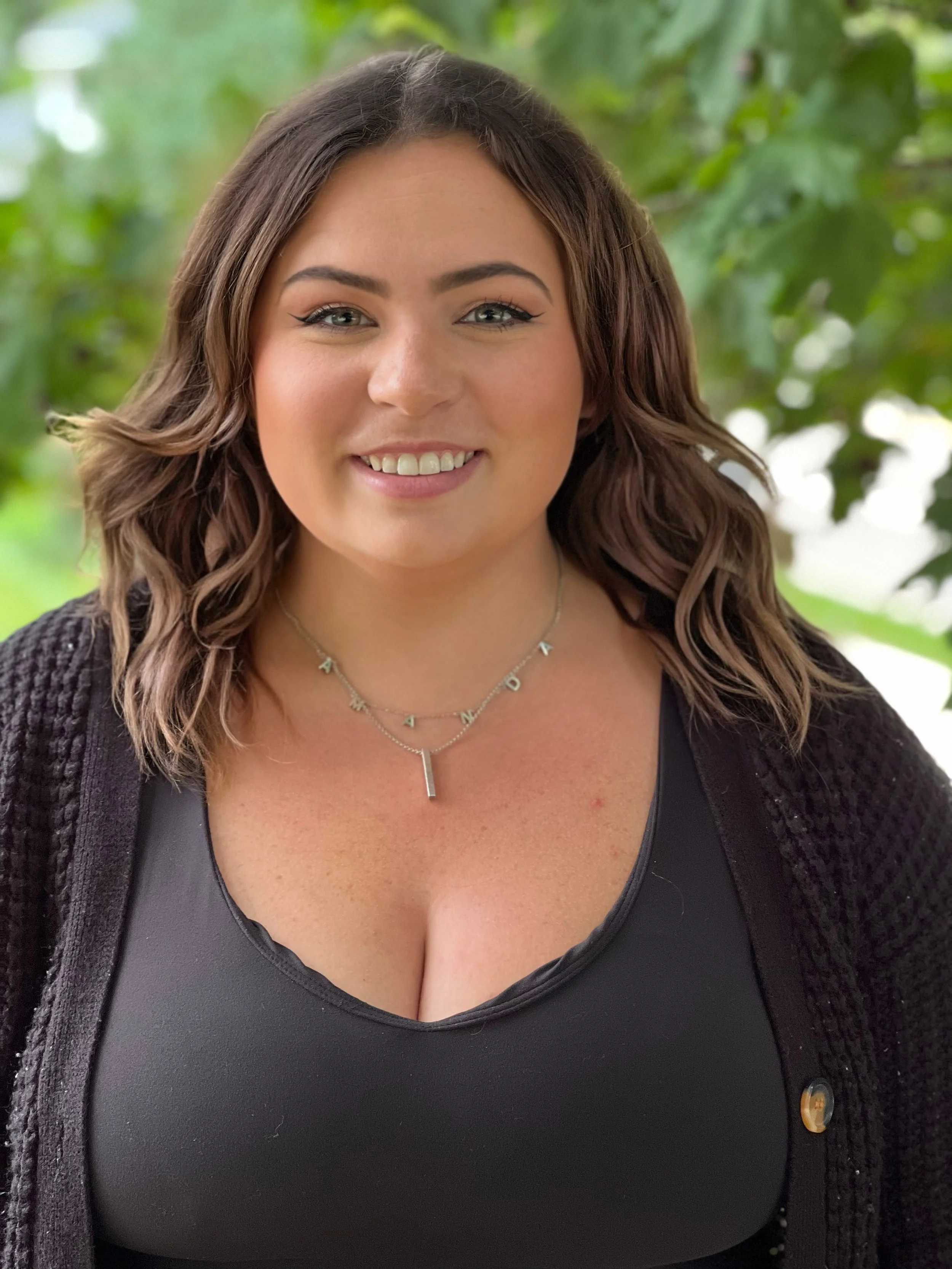 Smiling woman with wavy hair and black top, wearing a necklace, standing outdoors with green leaves in the background.