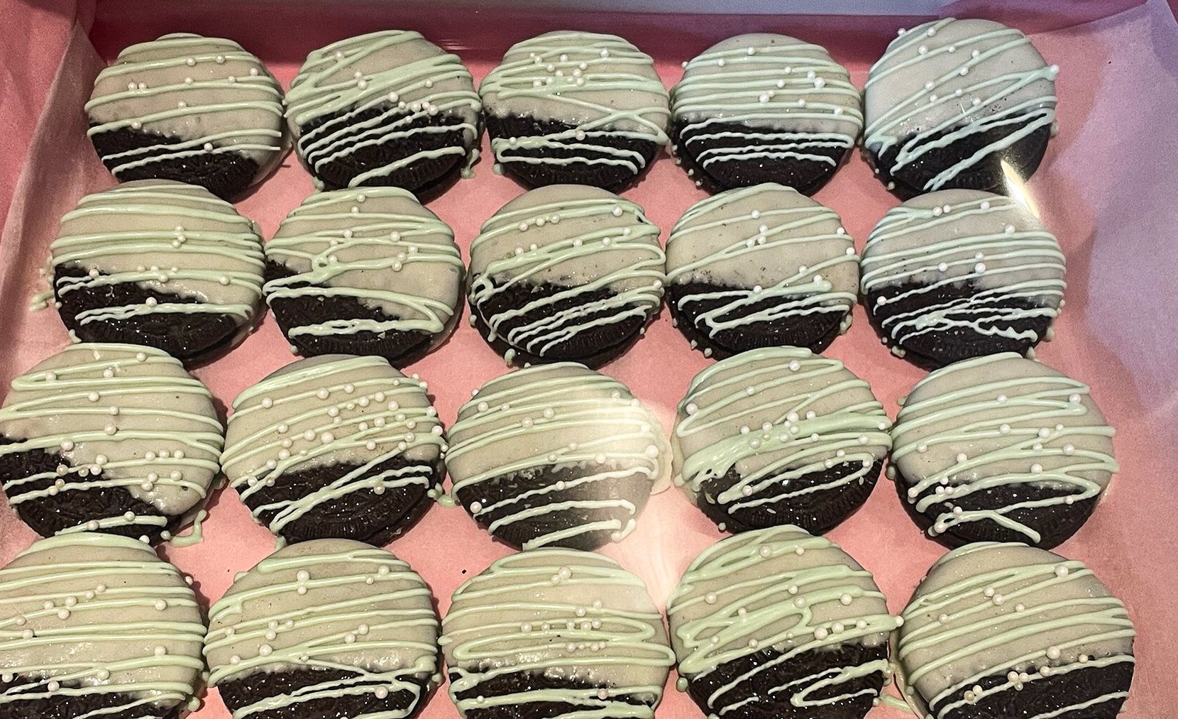 Chocolate cookies partially coated with white chocolate and drizzled with green icing, arranged in a pink box.