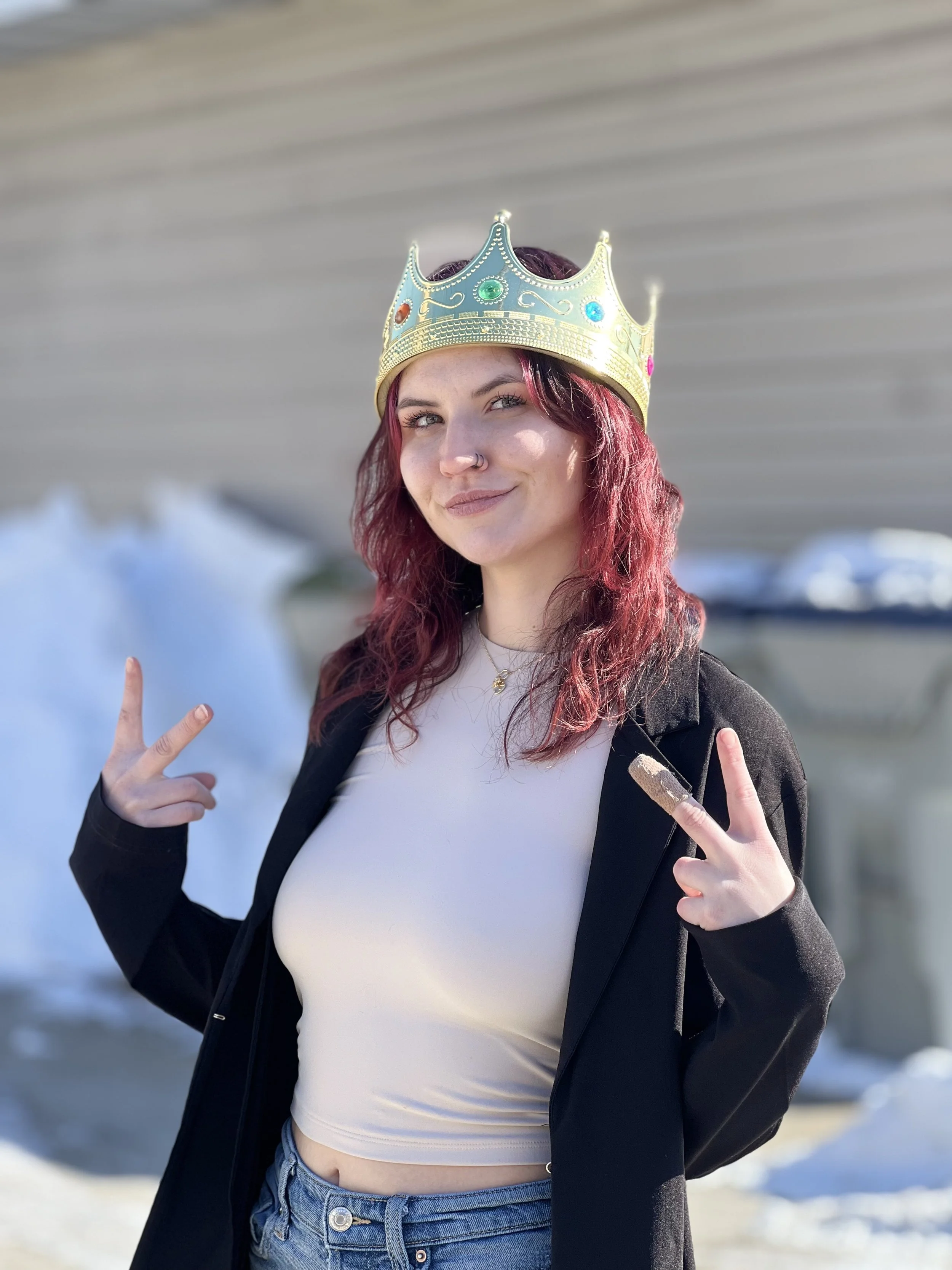 A person wearing a golden crown with colorful jewels stands outdoors. They have red hair and are making peace signs with both hands. They are dressed in a light crop top, dark jacket, and jeans, with snow in the background.