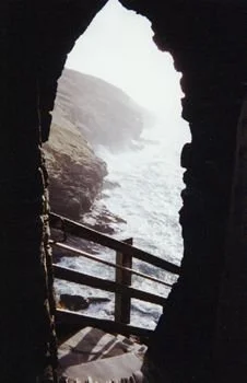 Vue depuis l'intérieur d'une grotte vers une mer agitée avec des falaises verdoyantes en arrière-plan, barrière en bois visible à l'entrée.