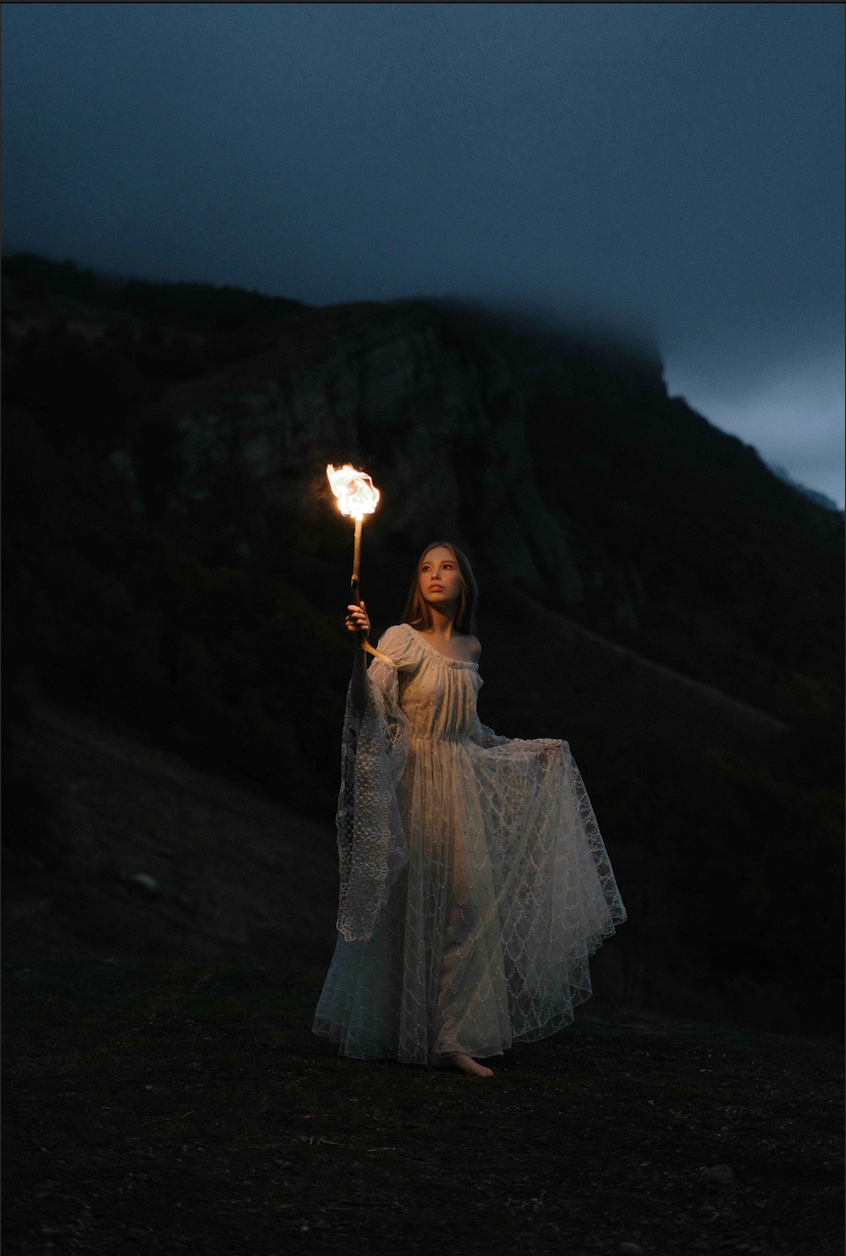 Femme vêtue d'une robe blanche portant une torche allumée, debout dans un paysage montagneux sombre.