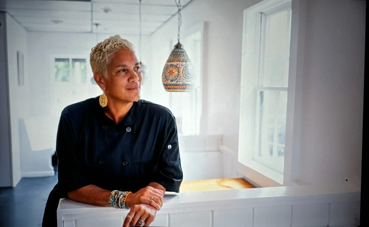 A woman with short blonde hair wearing a black chef's coat, sitting at a white counter with her hands folded, looking to her left. She has gold earrings and several bracelets on her right wrist. The background shows a brightly lit kitchen or dining area with white walls, windows, and a colorful hanging lamp.
