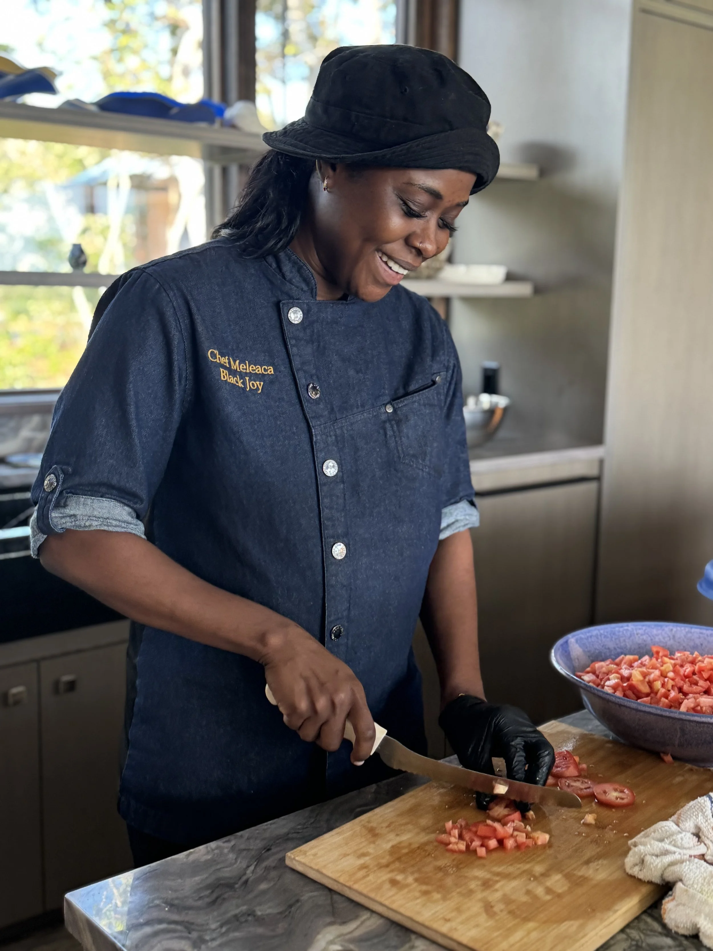 A woman wearing a black hat and a dark blue chef's coat is chopping tomatoes on a wooden cutting board in a kitchen.
