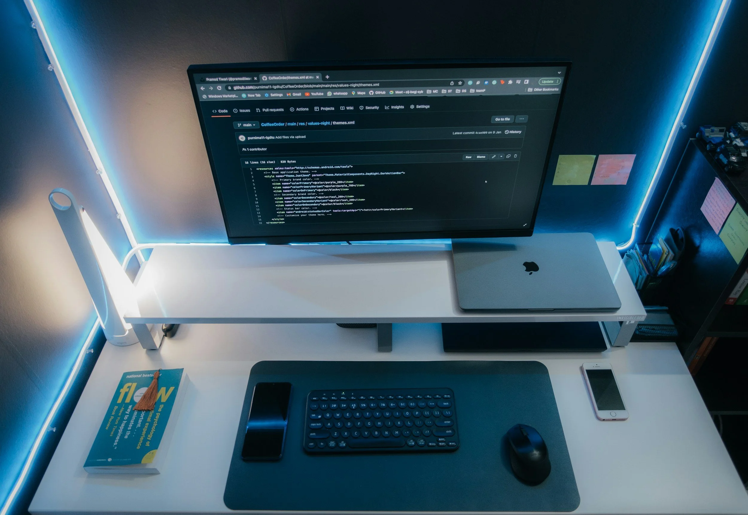 A testing desk setup with a large monitor showing code, a closed laptop, a smartphone, a wireless keyboard, a mouse on a desk mat, and a book titled 'Flow' on the left side of the desk. The desk is illuminated by LED strip lights around the edges and there are sticky notes on the wall behind.