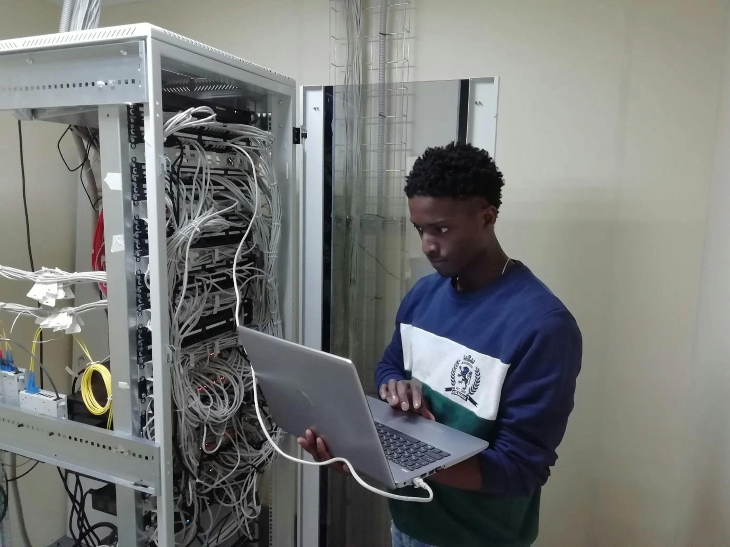 A young man working with a silver laptop in front of a server rack filled with tangled network cables.