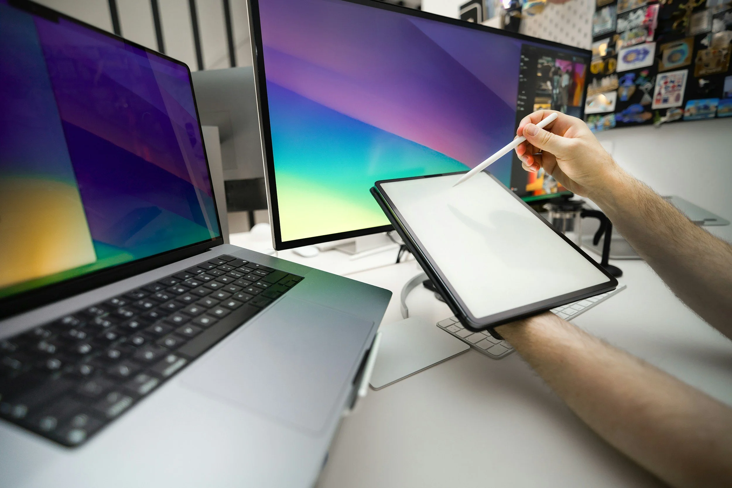 A person using a stylus on a graphics tablet, with multiple computer monitors displaying colorful backgrounds and a laptop nearby on a white desk.